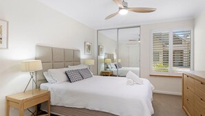 Ein Schlafzimmer mit Bett, Nachttischen, Deckenventilator, Fenster mit Jalousien und Spiegel.