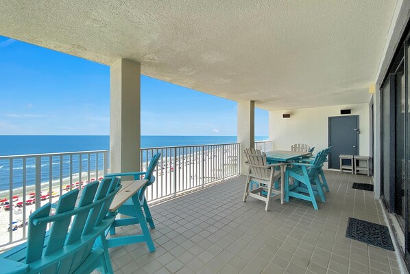 Un balcon avec des chaises turquoise et une table, donnant sur une plage parsemée de parasols et sous un ciel bleu azur.