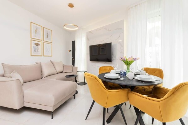 A modern living room with a beige sofa, a round dining table with yellow chairs, a flat-screen TV mounted on the wall, and a white floor.