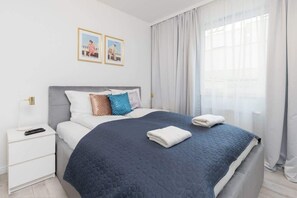 A neatly made bed with a dark quilt, a white bedside table with a remote control, and a window with sheer curtains.