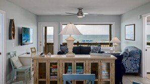 Un salon avec vue sur l’océan, un ventilateur de plafond, une table basse en bois et un canapé agrémenté de coussins à motifs.