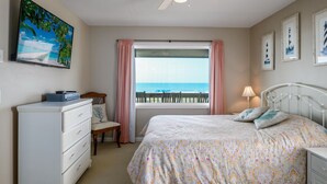 Une chambre à coucher comprenant un lit, une commode, une chaise, un ventilateur de plafond, une fenêtre donnant sur l’océan et des tableaux encadrés accrochés au mur.