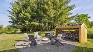 A circular outdoor seating area with four Adirondack chairs around a fire pit, a wooden pergola, and a stack of firewood. - The Greenhouse: Modern Rural Escape with Fire Pit (Ithaca)