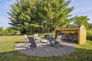 A circular outdoor seating area with four Adirondack chairs around a fire pit, a wooden pergola, and a stack of firewood. - The Greenhouse: Modern Rural Escape with Fire Pit (Ithaca)