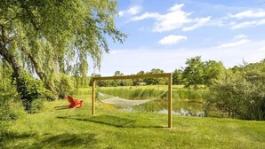 A hammock set up on a wooden frame in a grassy area with a pond and trees in the background. - The Greenhouse: Modern Rural Escape with Fire Pit (Ithaca)