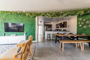 A modern living room with a green wall, a flat-screen TV, a sofa, and a dining area with a wooden table and chairs. - The Greenhouse: Modern Rural Escape with Fire Pit (Ithaca)