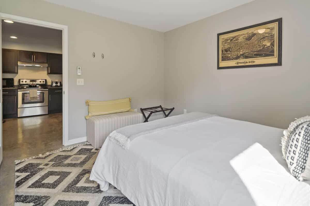 Une chambre avec un grand lit, une petite table de chevet et une chaise. On peut apercevoir une cuisine à travers une porte ouverte.