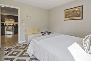 A bedroom with a large bed, a small bedside table, and a chair. There is a kitchen visible through an open door. - The Greenhouse: Modern Rural Escape with Fire Pit (Ithaca)