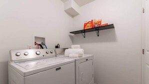 A laundry area with a front-loading washer and dryer, a shelf with laundry detergent, and a towel rack. - The Greenhouse: Modern Rural Escape with Fire Pit (Ithaca)