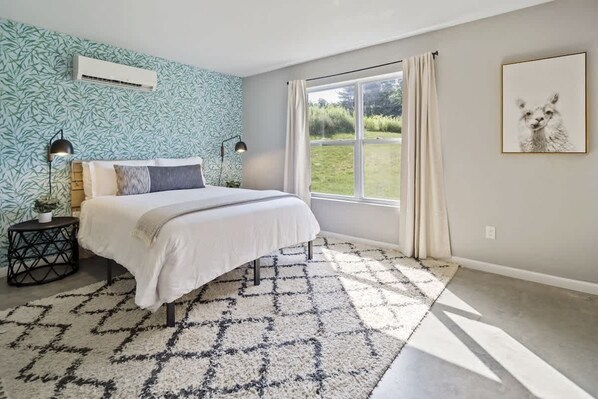 A bedroom with a large bed, a window with curtains, a wall-mounted air conditioner, and a framed picture on the wall. - The Greenhouse: Modern Rural Escape with Fire Pit (Ithaca)