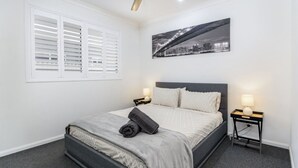 Un dormitorio ordenado con una cama, dos mesitas de noche, una mesita auxiliar, una ventana con contraventanas y un cuadro de un paisaje urbano en la pared.