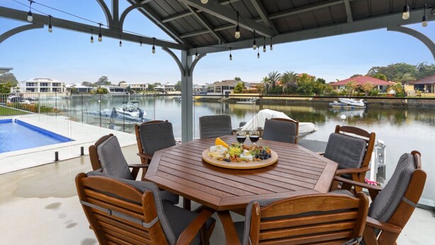 Una mesa de madera con sillas, una bandeja de frutas y copas de vino, con vistas a un puerto deportivo con barcos y casas.