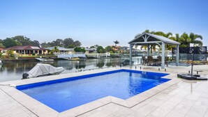 Una piscina con un área de estar techada, un muelle con barcos y una vista de casas y árboles al fondo.