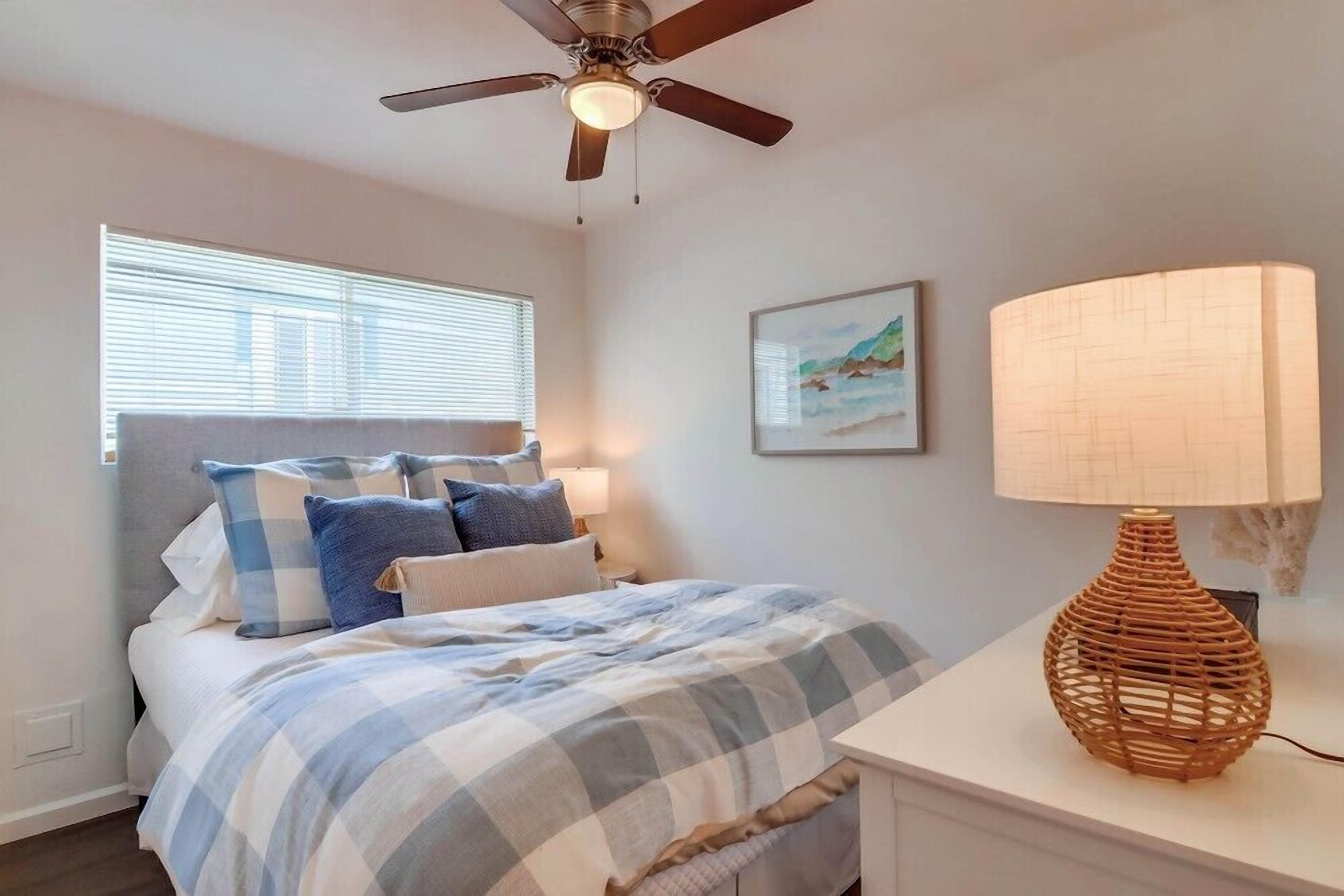 A bedroom with a bed, a ceiling fan, a bedside lamp, and a window with blinds.