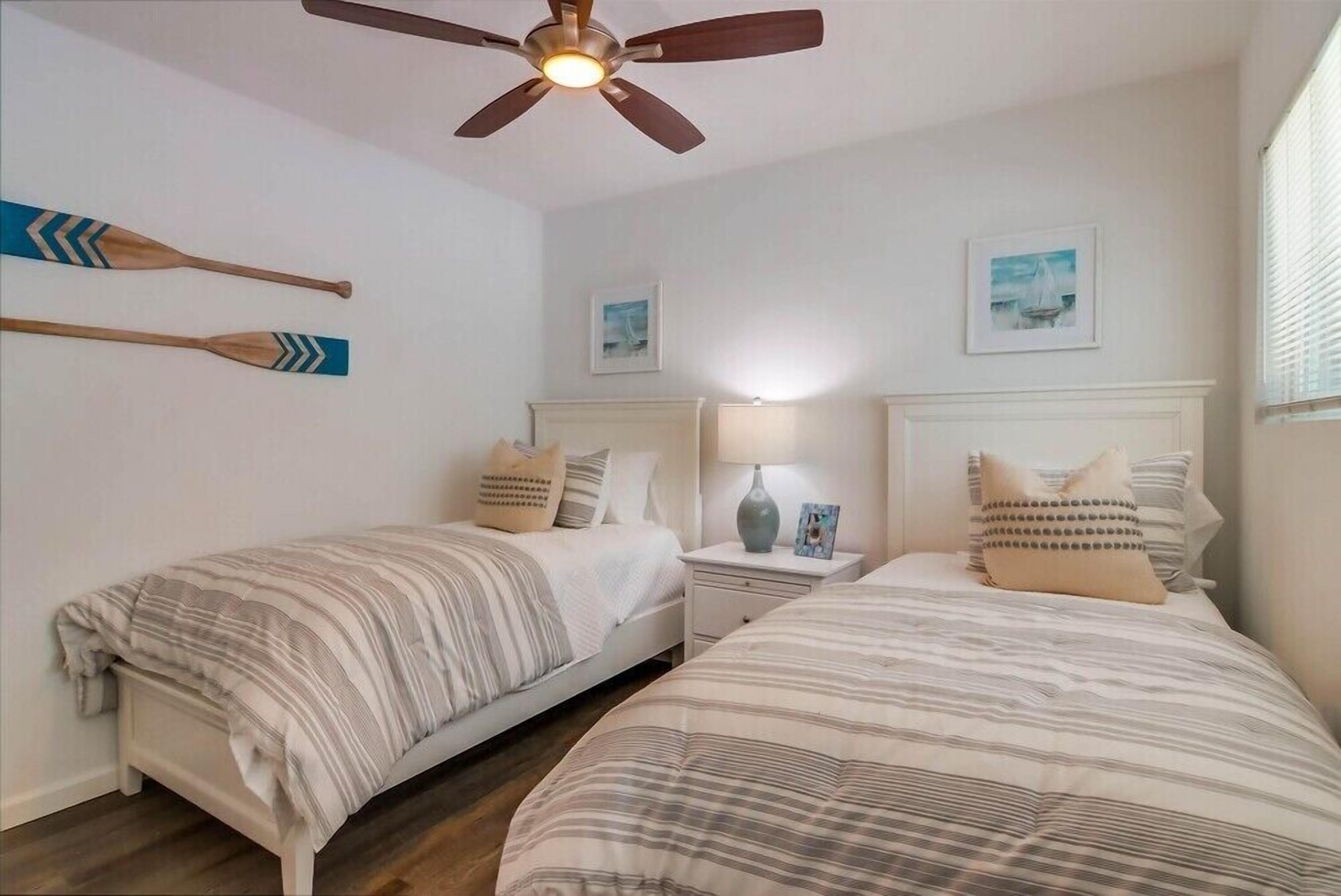 A bedroom with two single beds, a ceiling fan, bedside tables with lamps, and wall-mounted artwork.