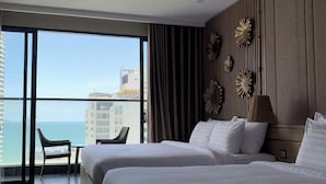 A hotel room with two beds, a large window with a view of the sea and buildings, a chair, and a decorative wall panel. - Virgo Hotel (Nha Trang)