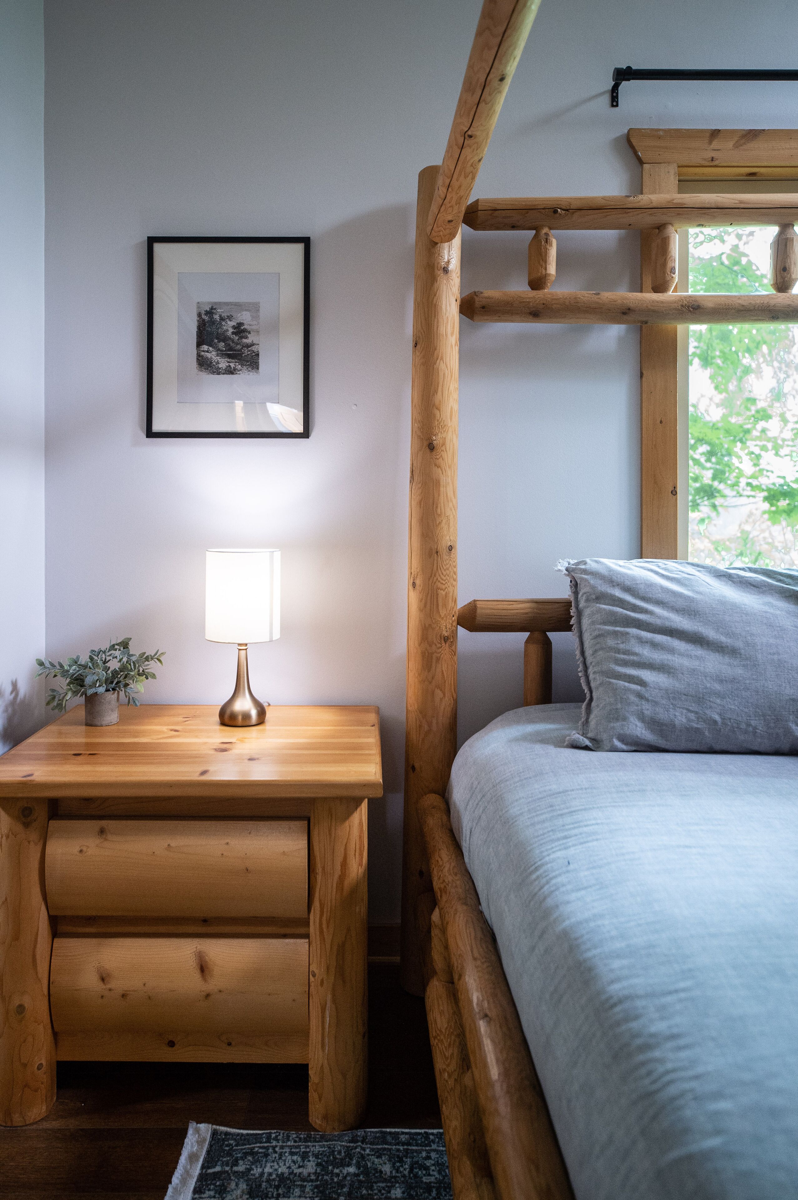 A wooden bunk bed with a grey pillow, a bedside table with a lamp, and a framed picture on the wall.