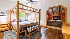 Una cama con dosel de madera, un banco, una chimenea de piedra y un ventilador de techo en una habitación con suelo de madera y ventana.