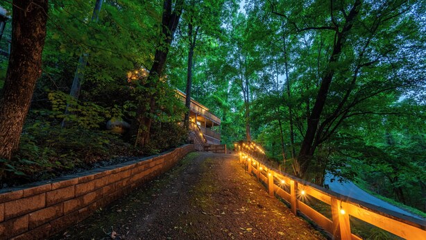Un camino que lleva a un edificio rodeado de árboles, con un muro de ladrillo a un lado y una valla de madera al otro, iluminado con luces colgantes.