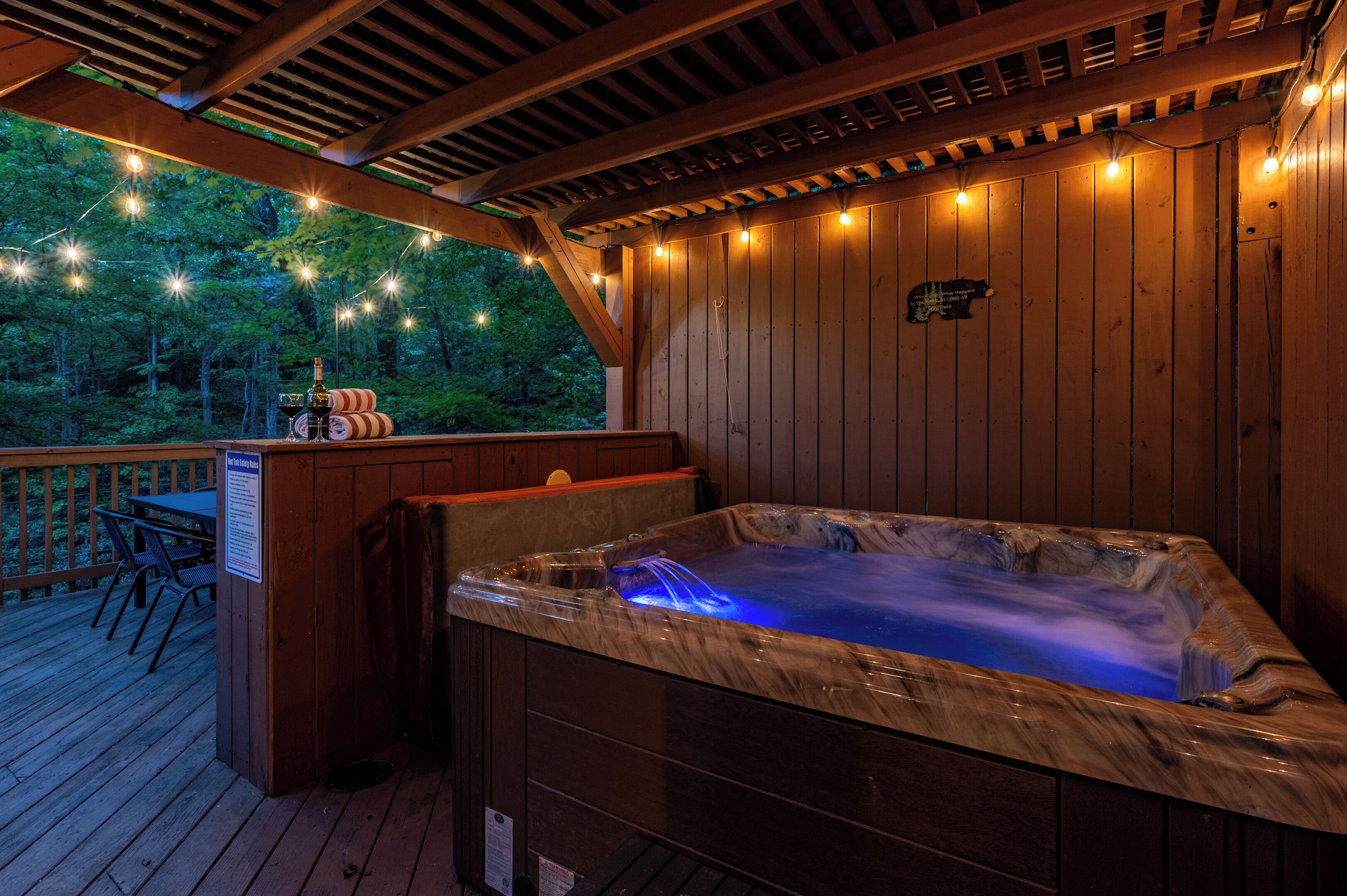 A hot tub on a wooden deck with string lights, a table with chairs, and a view of trees.
