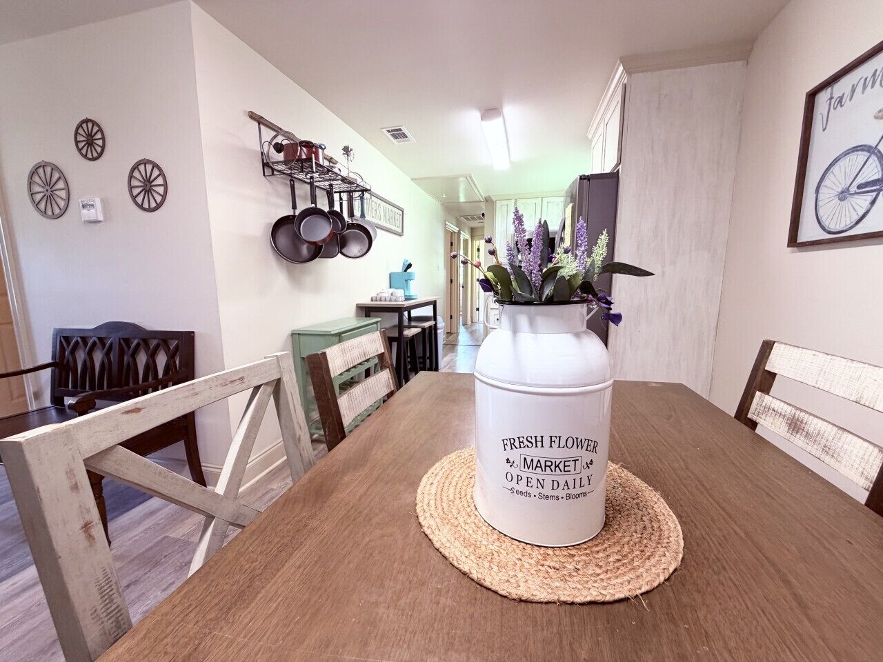 A dining table with a white container labeled 'FRESH FLOWER MARKET OPEN DAILY' and a vase with purple flowers.
