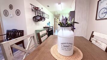 A dining table with a white container labeled 'FRESH FLOWER MARKET OPEN DAILY' and a vase with purple flowers.