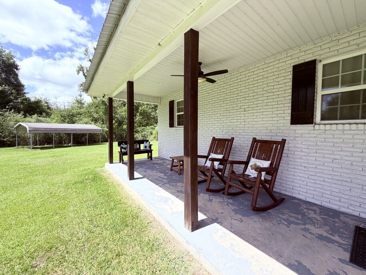 A covered porch with two rocking chairs and a table, a white brick building, and a grassy lawn.