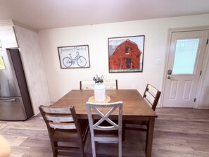 A dining area with a wooden table and chairs, a refrigerator, and two framed pictures on the wall.