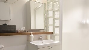 Une salle de bain avec un lavabo blanc, un miroir et une fenêtre à vitre dépolie. Il y a un tapis bleu sur le sol et une poubelle à côté du lavabo.