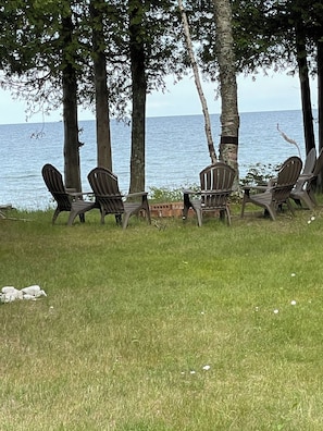 A row of Adirondack chairs facing a body of water, with trees in the background.