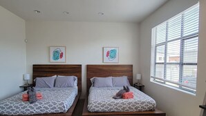 A hotel room with two beds, each with a wooden headboard, grey patterned bedding, and a window with blinds.