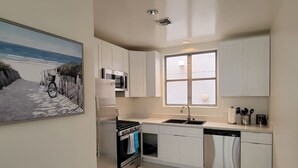 A modern kitchen with white cabinets, a built-in oven, microwave, and dishwasher. There's a window with a view of the beach and a painting of a beach scene on the wall.