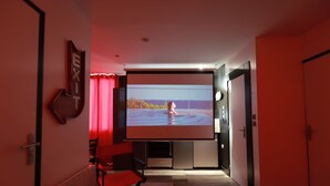 A modern hotel room with a large flat-screen TV mounted on the wall, a red curtain, a red chair, and a door. - Cinema apartment with home cinema and garage (Honfleur)