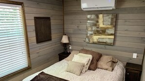 A bedroom with a bed, a nightstand, a lamp, a wall-mounted air conditioner, and a window with blinds. - Tiny Home only 1 mile to Vogel State Park!  Wandering Wood at the Reece Farm Vlg (Blairsville)