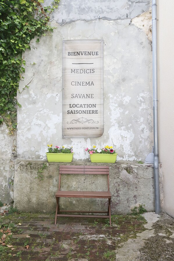 Een bord met de tekst 'BIENVENUE' en 'SAVANE LOCATION SAISONNIERE', twee bloempotten op een bankje en een verweerde muur.