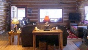 Hirsihirrestä rakennetun mökin sisustus: siellä on sohva, koriste-esineillä koristeltu pöytä, lamppu ja televisio.