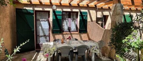 Un espace extérieur abrité, aménagé pour prendre les repas, avec une table et des chaises, entouré de murs en pierre et de volets verts.