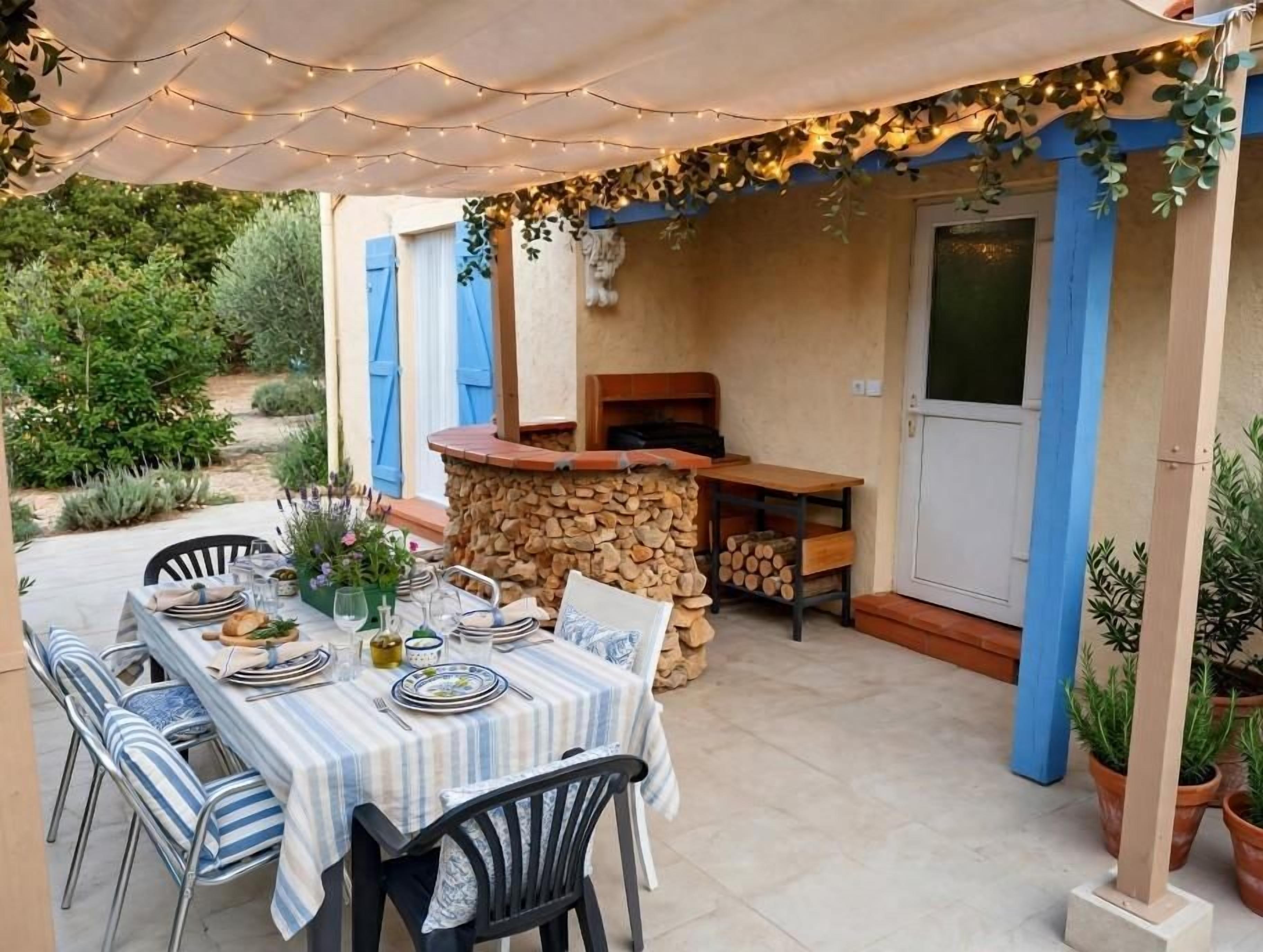 An outdoor dining area with a table set for two, surrounded by chairs, under a canopy with string lights.