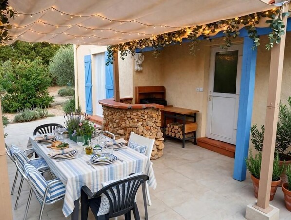 An outdoor dining area with a table set for two, surrounded by chairs, under a canopy with string lights.