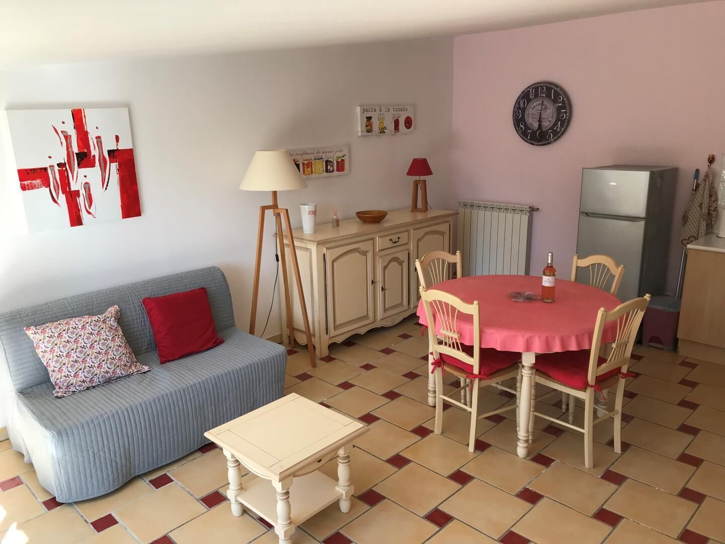 Een gezellige woonkamer met een blauwe bank, een klein tafeltje, een eettafel met stoelen, een lamp, een klok en een koelkast.