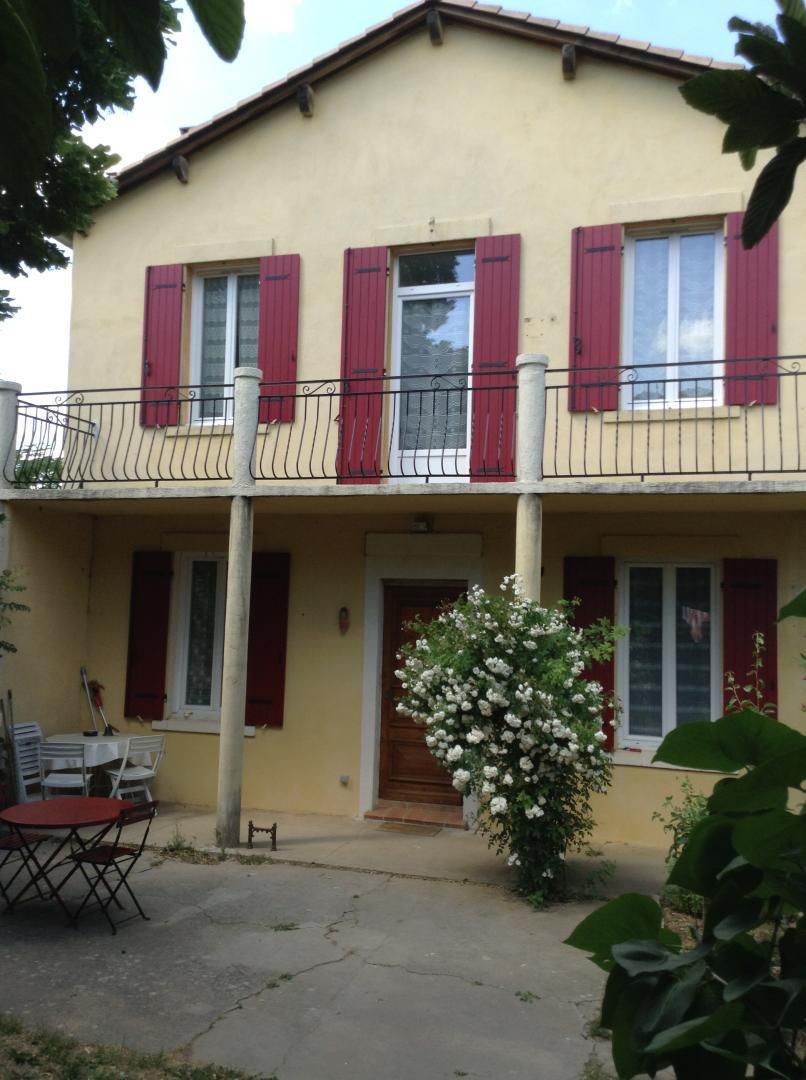 A two-story building with red shutters, a balcony with a wrought iron railing, and a small garden area with a white flowering plant.