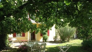 An outdoor seating area with a table and chairs under a tree, a yellow building with red shutters in the background, and a grassy lawn with flowers.