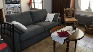 A living room with a grey sofa, a wooden table, and a chair. There is a window with curtains, a door, and a kitchen in the background.