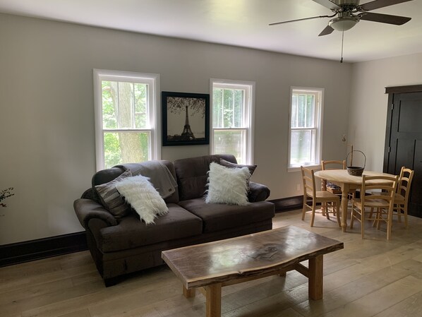 A living room with a brown sofa, a wooden coffee table, and a dining table with chairs. There are three windows providing natural light. - 3-bedroom vacation home just 20 minutes from Six Flags St. Louis (Ballwin)