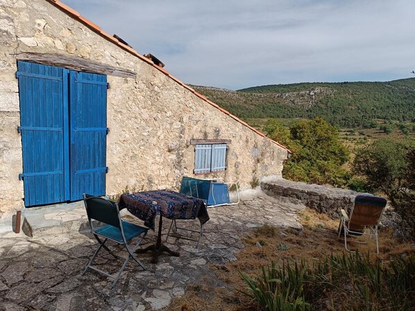 A rustic stone building with blue shutters, a small table with a patterned tablecloth, and two chairs. - Le Gîte Panda Saint-maymes (TRIGANCE)