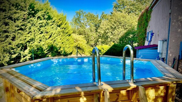 A wooden hot tub with two metal ladders, surrounded by lush greenery and a clear blue sky.