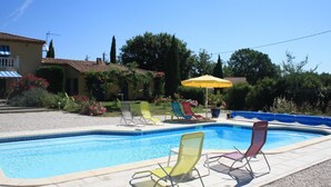 Una piscina con sillones de descanso y una sombrilla, rodeada de un jardín y un edificio con tejado de baldosas.