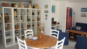 Una sala de estar con una mesa de comedor de madera, sillas blancas, un sofá azul y un estante blanco lleno de diversos objetos decorativos.