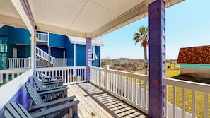 A wooden deck with a white railing, a staircase leading to a blue building with white trim, and a view of the beach and ocean.