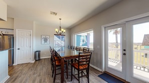 A dining area with a large table and chairs, a trash can, a refrigerator, and a door leading to a balcony with a view of the beach.
